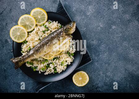 Deliziosa trota arrosto condita con spezie e succo di limone e accompagnata da gustoso riso bianco con piselli. Servito su un elegante piatto nero. Spazio per Foto Stock