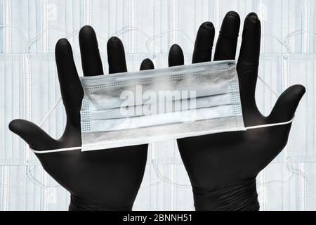 Maschera protettiva per il viso paramedico in due guanti medici neri in mani sullo sfondo di bende respiratorie per il viso umano Foto Stock