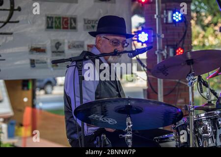 Senior maschio, vestito con abiti acutamente, con fedora Hat, suonando batteria, in rock band al concerto di outoor. Foto Stock