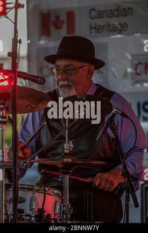 Senior maschio, vestito con abiti acutamente, con fedora Hat, suonando batteria, in rock band al concerto di outoor. Foto Stock