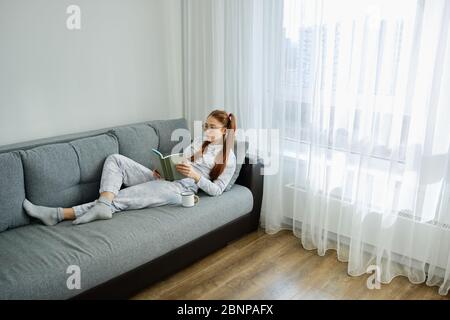 Ragazza rossa in pigiama e bicchieri si trova sul divano e legge un libro, cornice da sopra Foto Stock
