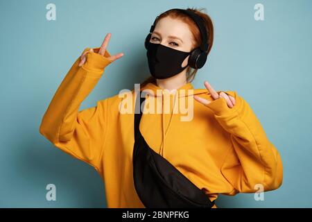 Una ragazza dai capelli rossi con cuffie, felpa con cappuccio gialla, maschera protettiva, si alza e mostra il marchio del mondo con le mani Foto Stock