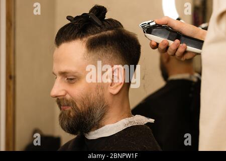 Rasoio elettrico per taglio di capelli maschile. La mano del parrucchiere rasa i capelli con il rasoio elettrico. Hipster man ottenere un taglio di capelli trendy con un regolacapelli e pettine. Servizi Barbiere. Procedura di acconciatura. Foto Stock