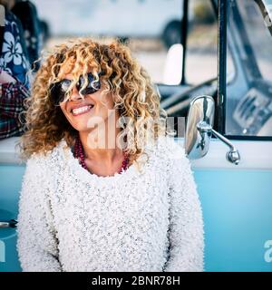 Cheful e bella bionda caucasica adulta giovane donna sorridente e ridente con occhiali da sole e vintage van blu in background - concetto di gente felice Foto Stock