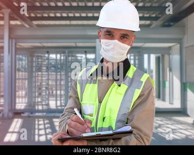 Operaio di costruzione in hardhat bianco e mascherine protettive con clipboard davanti checkpoint con tornello su sfondo. Foto Stock