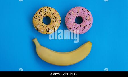 Due ciambelle e una banana giacciono sulla superficie blu, formando un'astrazione dell'emoticon - sorriso. Preso dal punto più alto Foto Stock