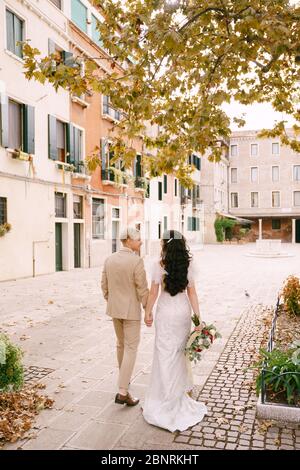 Matrimonio in Italia a Venezia. Sposa e sposo camminano lungo le strade deserte della città. Gli sposi novelli stanno tenendo la mano sotto l'albero con il fogliame giallo dell'autunno Foto Stock