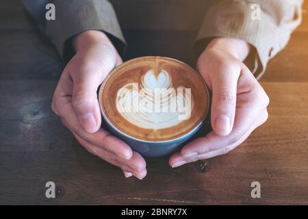 Immagine closeup delle mani che tengono una tazza blu di caffè latte caldo con l'arte del latte su un tavolo di legno nel caffè Foto Stock