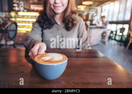 Immagine closeup di una donna asiatica che tiene una tazza blu di caffè latte caldo con latte art su tavolo di legno nel caffè Foto Stock