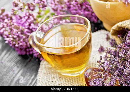 Tisana origano in una tazza di vetro su tovagliolo di burlap, fiori di maggiorana essiccati in sacchetto e cucchiaio, fiori freschi in un mortaio e sul tavolo contro il legnoso scuro Foto Stock