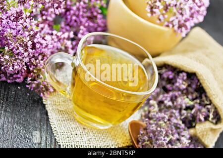 Tisana origano in una tazza di vetro su tovagliolo di burlap, fiori freschi in mortaio e sul tavolo, fiori di maggiorana secchi in una borsa e cucchiaio su legno scuro b Foto Stock