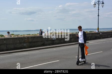 Un pilota di scooter elettronico guarda il suo cellulare mentre si guida sulla strada chiusa sul lungomare di Southsea, Hampshire, come il pubblico è stato invitato a non affollarsi nelle città costiere e nei parchi nazionali in tutta l'Inghilterra durante il clima caldo. Foto Stock