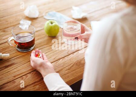 Angolo alto di donna anonima con pillole e tazza d'acqua che strega a tavola con tè e mela durante la malattia Foto Stock