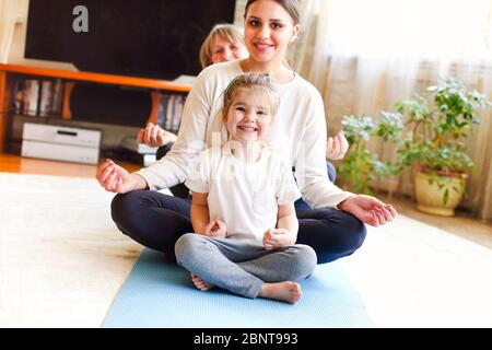 Donne adulte e bambina che si siedono in linea sul pavimento e si allungano le braccia mentre si fa yoga a casa insieme Foto Stock