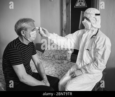 Istantanea in bianco e nero dell'esame medico a casa del paziente durante la quarantena. Mani del medico che misura la febbre di un paziente anziano maschile con un termometro a infrarossi. Cartello di rischio biologico sullo sfondo Foto Stock