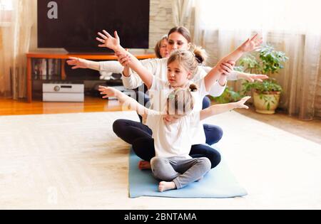 Donne adulte e bambine che si siedono in fila sul pavimento e si stendono le braccia mentre si fa yoga a casa insieme Foto Stock