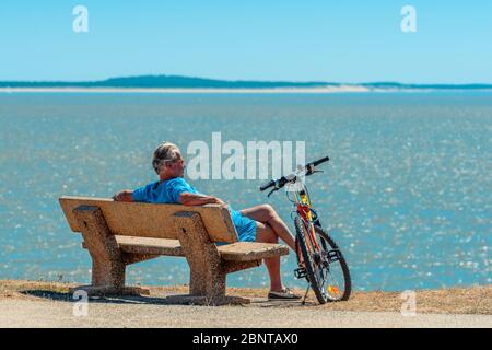 Saint-Palais-sur-Mer, Francia: Un ciclista si siede su una panchina con la sua bicicletta al suo fianco guardando il mare - la baia di Biscay, un golfo dell'Oceano Atlantico. Foto Stock