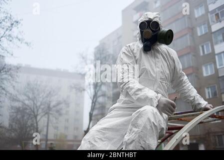 Ambientalista maschile in uniforme protettiva che tiene le pinzette mentre si riposa su un terreno sportivo di strada. Lo scienziato indossa tuta per radiazioni, maschera a gas e guanti sterili. Concetto di ricerca ed ecologia. Foto Stock