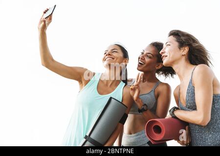 Foto di tre multietnica atletico sportivi in tute sorridente e tenendo selfie sul cellulare mentre in piedi oltre il muro bianco all'aperto Foto Stock