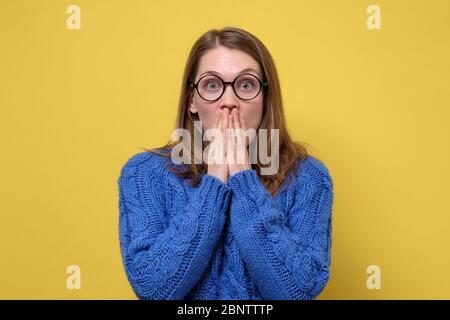 Donna emozionata sorpresa in pullover blu e occhiali che la ricopre la bocca con le mani in shock. Ha perso la voce. Emozione facciale umana sulla parete gialla. Foto Stock