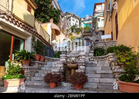 Colorata stradina con fontana e fiori vicino a Piazza Duomo a Taormina in sole mattina, Sicilia, Italia Foto Stock