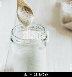 Pronto per preparare il pane di pasta di grano soursough antipasto gocciolando da cucchiaio di legno. Primo piano della vista del vaso di vetro con base di pasta acida idratazione più alta su sfondo di legno bianco. Concetto di panificazione fatto in casa Foto Stock