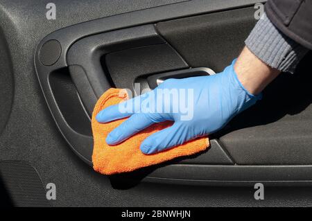 La mano del conducente in un guanto di protezione blu strofina con un panno una maniglia interna della portiera della vettura. Coronavirus o Covid-19 disinfezione auto Foto Stock