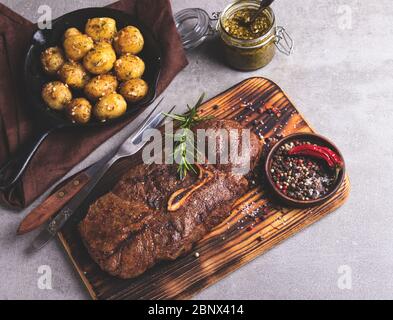 bistecca di manzo alla griglia con osso su un tagliere, patate arrosto in una padella Foto Stock