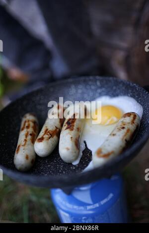 Preparare la salsiccia d'uovo su una padella con una stufa a gas da campeggio al campo esterno usando un bastone di legno come utensile da cucina. Foto Stock