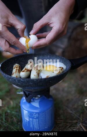 Preparare la salsiccia d'uovo su una padella con una stufa a gas da campeggio al campo esterno usando un bastone di legno come utensile da cucina. Foto Stock