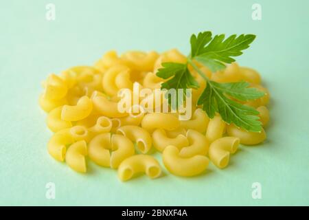 Un mucchio di piccoli corni di maccheroni crudi e prezzemolo fresco su sfondo verde pastello. Pasta di grano duro. Ingrediente per mangiare l'healhy e cibo vegetariano. Foto Stock
