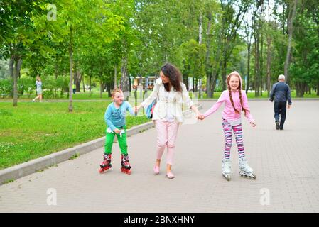 Una giovane madre rotola le mani di bambini allegri su pattini a rotelle in un Sunny Park in estate a San Pietroburgo Foto Stock