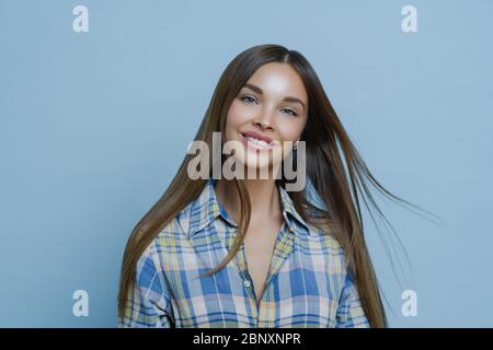 Affascinante giovane donna sorride stuzzicante, vestita in camicia a scacchi, ha capelli lunghi e dritti scuri, fa foto per profilo personale in rete sociale Foto Stock