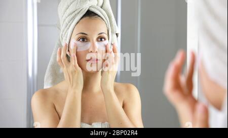 Donna di bellezza che applica la maschera sotto-occhio di anti-affaticamento guardando se stessa nello specchio in bagno. Cura della pelle ragazza toccare le macchie di tessuto maschera sotto gli occhi a. Foto Stock