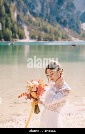 Bella sposa in abito bianco con maniche e pizzo, con bouquet giallo autunnale di fiori secchi e rose di pony, sul Lago di Braies in Italia Foto Stock