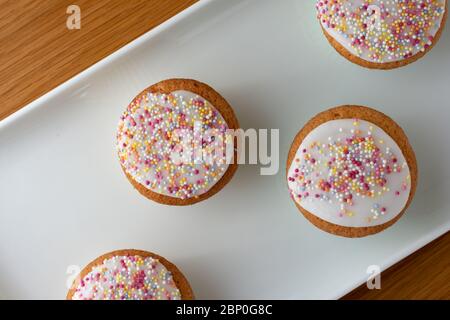 Quattro torte ghiacciate su un vassoio bianco, vista dall'alto verso il basso e ricoperte da spruzzi di zucchero multicolore. Il vassoio bianco è posizionato sopra a. Foto Stock