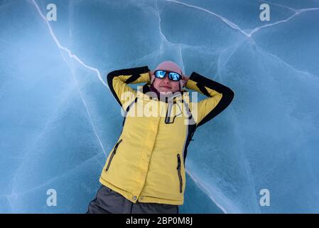 Ragazza felice in giacca nera gialla, pantaloni grigi, occhiali da sole blu a specchio e cappello rosa giace sul ghiaccio blu cracked del lago Baikal. Vista dall'alto Foto Stock