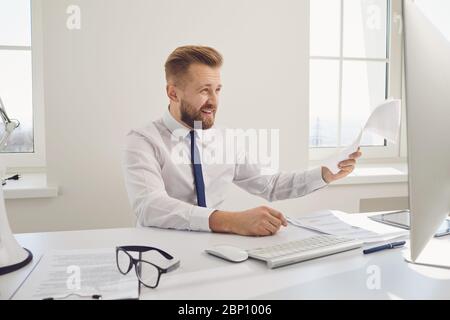 Serio uomo d'affari occupato documenti di lavoro seduto ad un tavolo con un computer in ufficio. Foto Stock