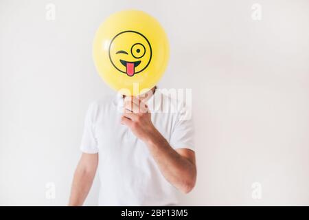 Giovane isolato su sfondo bianco. Ragazzo in camicia tenere palloncino giallo in faccia anteriore. Emoji dipinte con lingua esterna. Nascondere o coprire la faccia Foto Stock