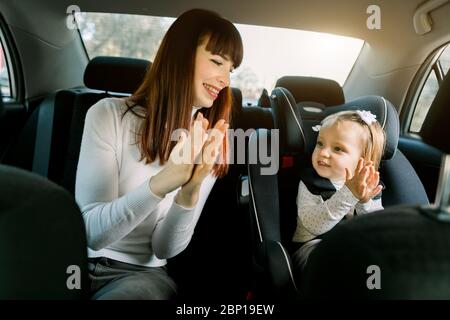 Bella donna ridente, giovane madre seduta in un'automobile, avendo divertimento con la sua bambina in un seggiolino per bambini, aggrappando le mani e sorridendo. Bambino Foto Stock