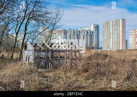 Su un vecchio campo abbandonato c'è una serra distrutta per le verdure sullo sfondo di edifici a più piani in costruzione Foto Stock