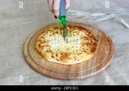 La mano della ragazza taglia un coltello khachapuri con un coltello in pezzi su una tavola da cucina su uno sfondo di burlap Foto Stock