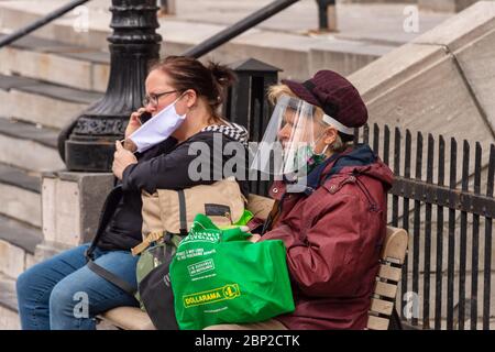 Montreal, CA - 16 maggio 2020: Due donne che indossano maschera facciale e scudo di protezione del viso seduti su una panca. Foto Stock