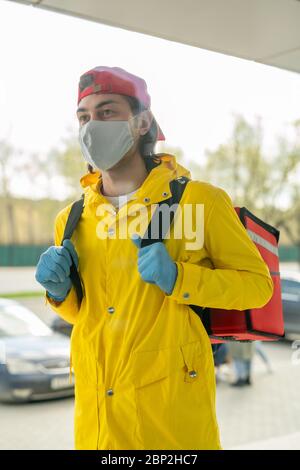 Giovane uomo in gialle da lavoro, cappellino, guanti protettivi e maschera che va a uno dei clienti mentre trasportano grande zaino rosso con ordini Foto Stock