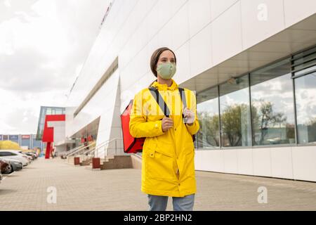 Carino giovane corriere in trench giallo e maschera protettiva che cammina lungo il supermercato contemporaneo mentre si precipitano ai clienti Foto Stock