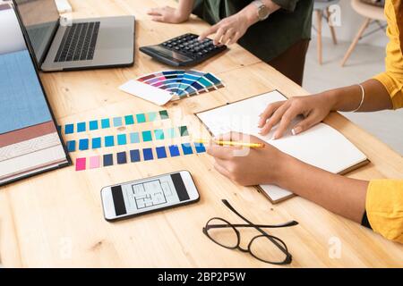 Giovane designer con matita che punta ai campioni di colore mentre fa note sullo sfondo dei colleghi che premono i pulsanti della calcolatrice Foto Stock