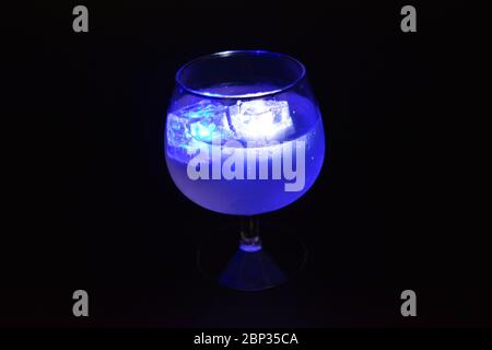 Blu e bianco cubetti di ghiaccio luminoso che galleggiano in una deliziosa bevanda fredda bianca ghiacciata si trovano in un bicchiere cognac su uno sfondo nero smerigliato Foto Stock
