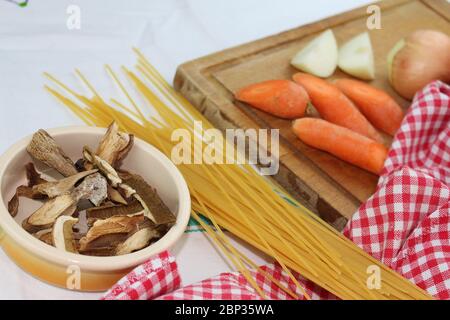 Prodotti alimentari disposti sul tavolo con volant, con quadrati rossi e bianchi. Cucina mediterranea italiana con spaghetti ai funghi porcini secchi. Foto Stock