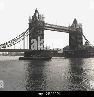 Anni '50, storica, una chiatta a vapore dell'epoca che passa sotto il Tower Bridge, un famoso punto di riferimento londinese, con Butler's Wharf sul lato sud del fiume Tamigi. Un ponte combinato tra il 1886 e il 1894, costruito tra il Tamigi, il ponte attraversa il Tamigi vicino alla Torre di Londra ed è diventato un simbolo iconico della città. Foto Stock