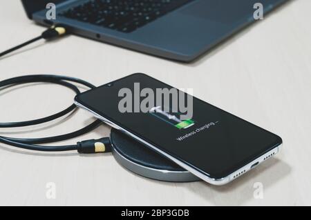 Gli smartphone moderni si ricaricano tramite il caricabatterie wireless situato accanto al computer portatile su un tavolo di legno. Concetto tecnologico moderno. Messa a fuoco selettiva Foto Stock